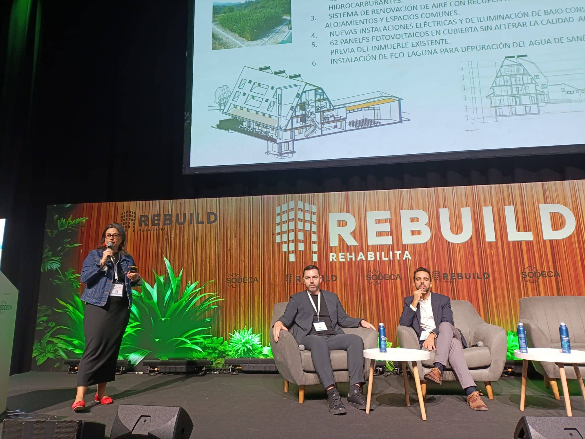 María San Juan, de pie con micrófono, presenta el proyecto Co-Rural Robregordo Senior Cohousing en el Congreso Nacional de Rehabilitación Avanzada. Detrás, una pantalla muestra planos y puntos clave del proyecto. A su lado, dos hombres sentados escuchan atentamente en un escenario decorado con un cartel de Rebuild Rehabilita y plantas en el fondo