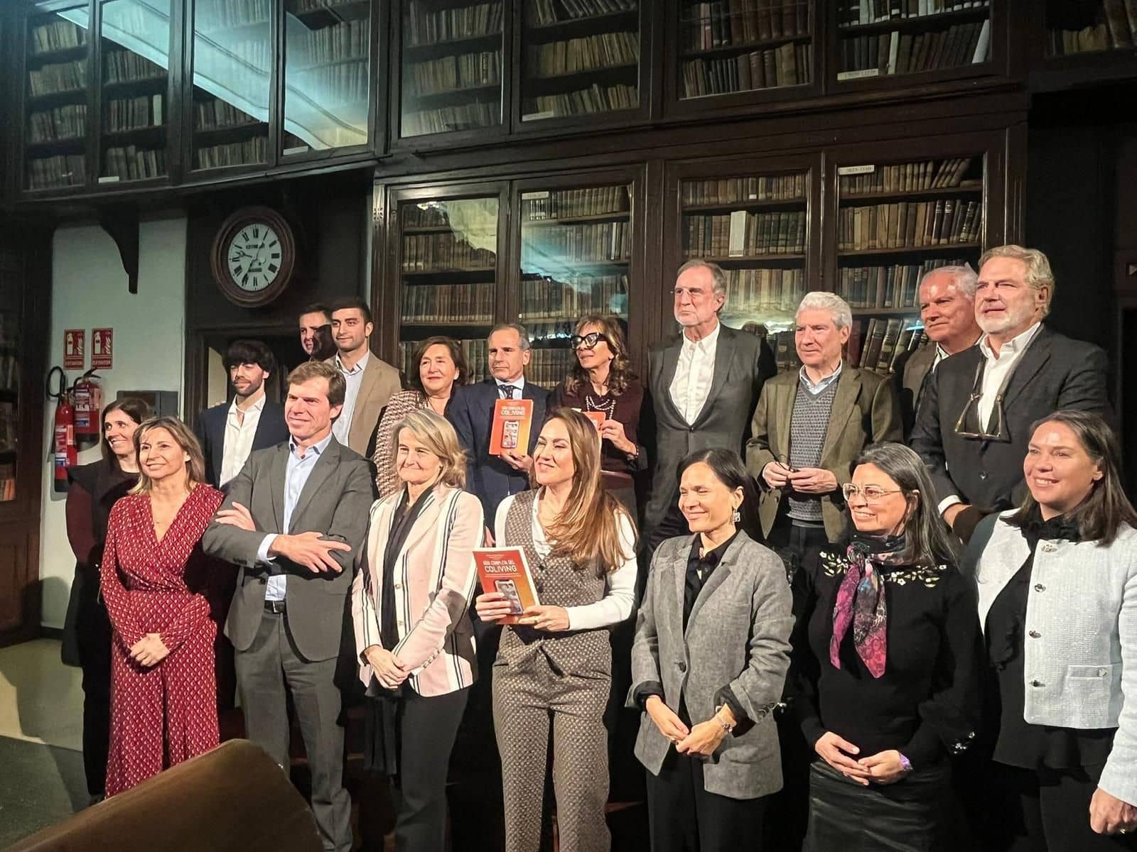 La imagen muestra a diecisiete personas de pie en una librería, posando en grupo frente a una estantería llena de libros. Varias personas sostienen en sus manos el libro "Guía completa de Coliving".