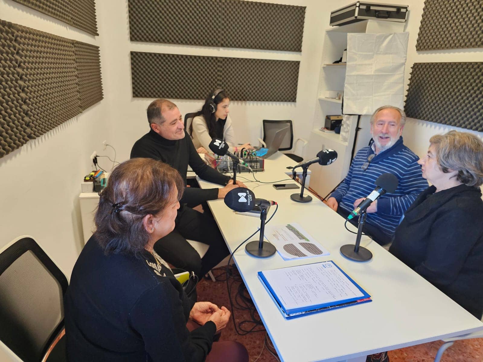 La imagen muestra al presidente de Hábitat Colaborativo y dos personas socias del Residencial Josefina Castro en una sala de grabación, sentadas alrededor de una mesa blanca con micrófonos. En el fondo, se observan paneles de insonorización en las paredes. Una persona está frente a una consola de audio.