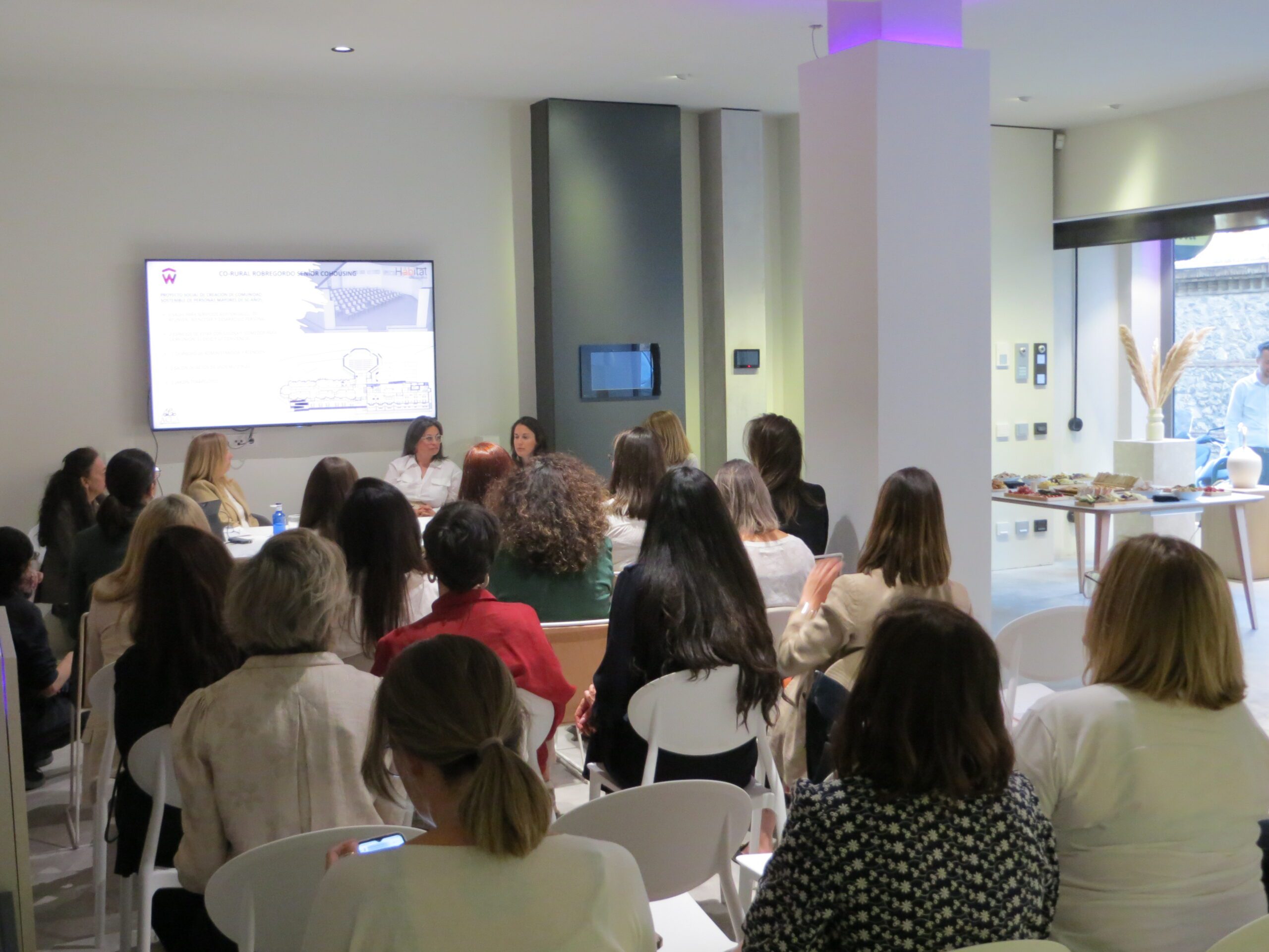 La imagen muestra un salón de actos con un grupo de más de 15 mujeres sentadas de espaladas, mirando hacia una pantalla donde se muestra una imagen del proyecto Corural Robregordo Senior Cohousing. Al fondo se ve cuatro ponentes mujeres, también sentadas.