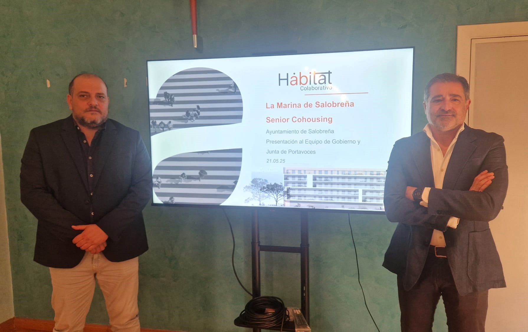 La imagen muestra a dos hombres de pie frente a una pantalla de televisión que presenta un proyecto llamado "La Marina de Salobreña Senior Cohousing". El hombre de la izquierda (Javier Ortega Prados )lleva una chaqueta oscura y pantalones claros; el hombre de la derecha (José Carlos Rodrigo) viste una chaqueta azul y pantalones oscuros y tiene los brazos cruzados.