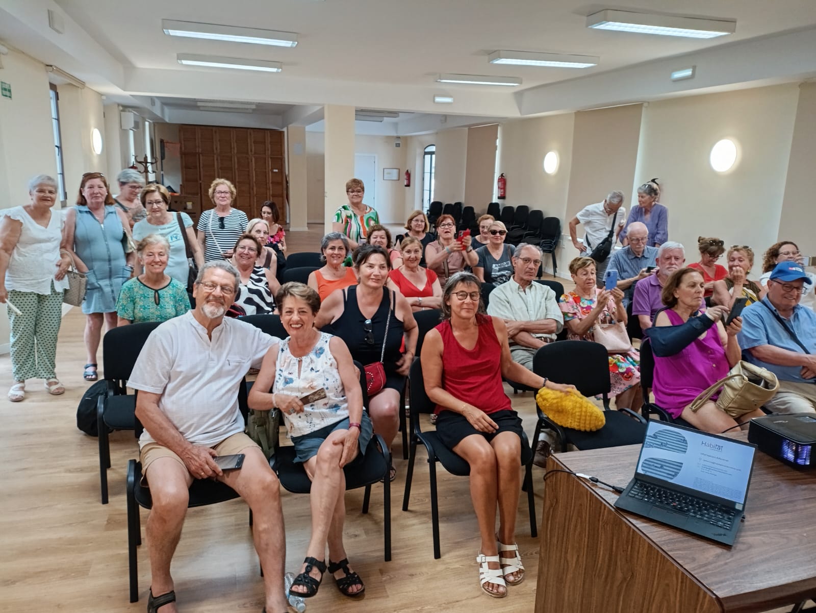 Grupo de unas 20 personas sentadas en filas frente a una mesa central y sonriendo a la cámara, durante la presentación del proyecto La Marina de Salobreña Senior Cohousing